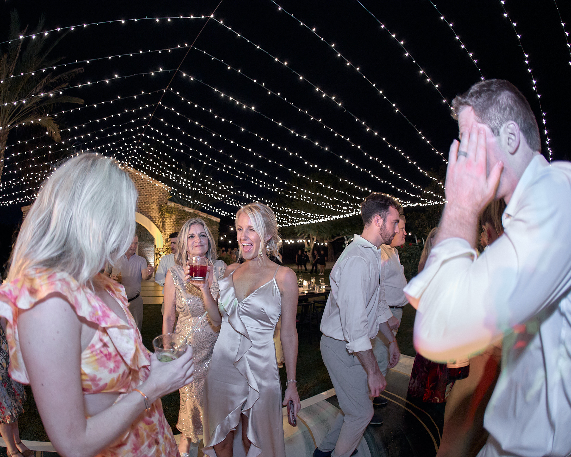 A group of people dancing under string lights at a wedding.