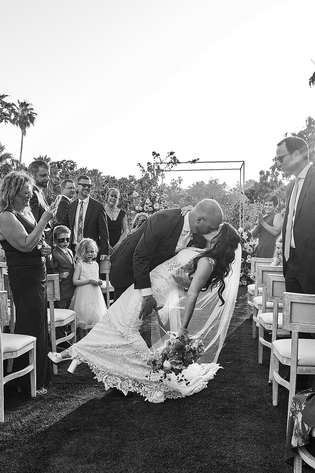 A newlywed couple shares a kiss in a dip pose as guests clap and smile around them during an outdoor wedding ceremony. A newlywed couple shares a kiss in a dip pose as guests clap and smile around them during an outdoor wedding ceremony.